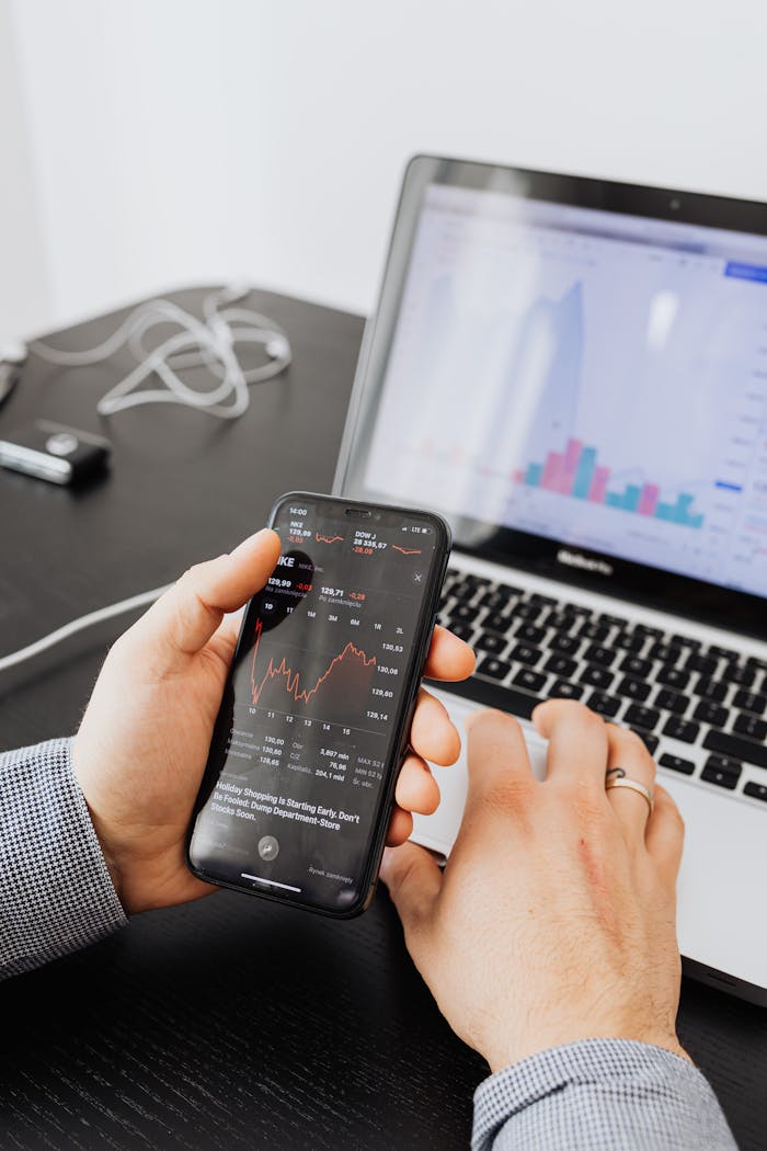 Hands holding smartphone analyzing stock market data next to laptop displaying charts.